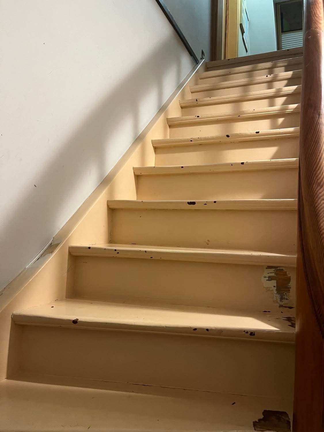 Drewniane schody w kolorze kremowym z widocznymi śladami użytkowania i ubytkami farby, widok z dołu na górę, z fragmentem drewnianej poręczy po prawej stronie.