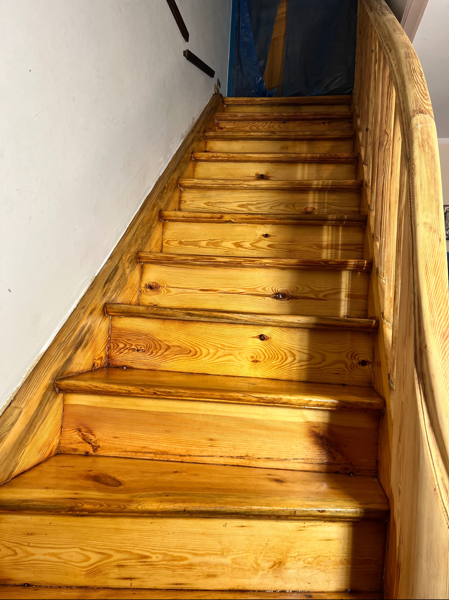 Drewniane schody po renowacji, widoczne świeże lakierowanie i odnowiona balustrada, fragment ściany z widocznymi śladami po remoncie.