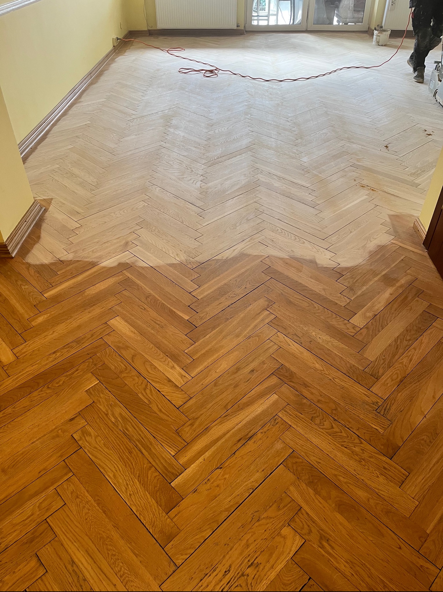 Cyklinowanie parkietu w jodełkę: widoczna różnica między obszarem po cyklinowaniu a obszarem przed obróbką, z maszyną cykliniarską w tle.