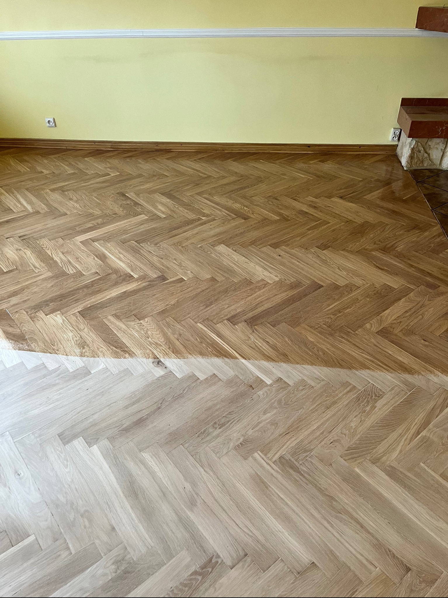 Parkiet ułożony w jodełkę, w połowie wycyklinowany, ukazujący różnicę w kolorze i fakturze przed i po zabiegu, ściana w tle w kolorze żółtym.