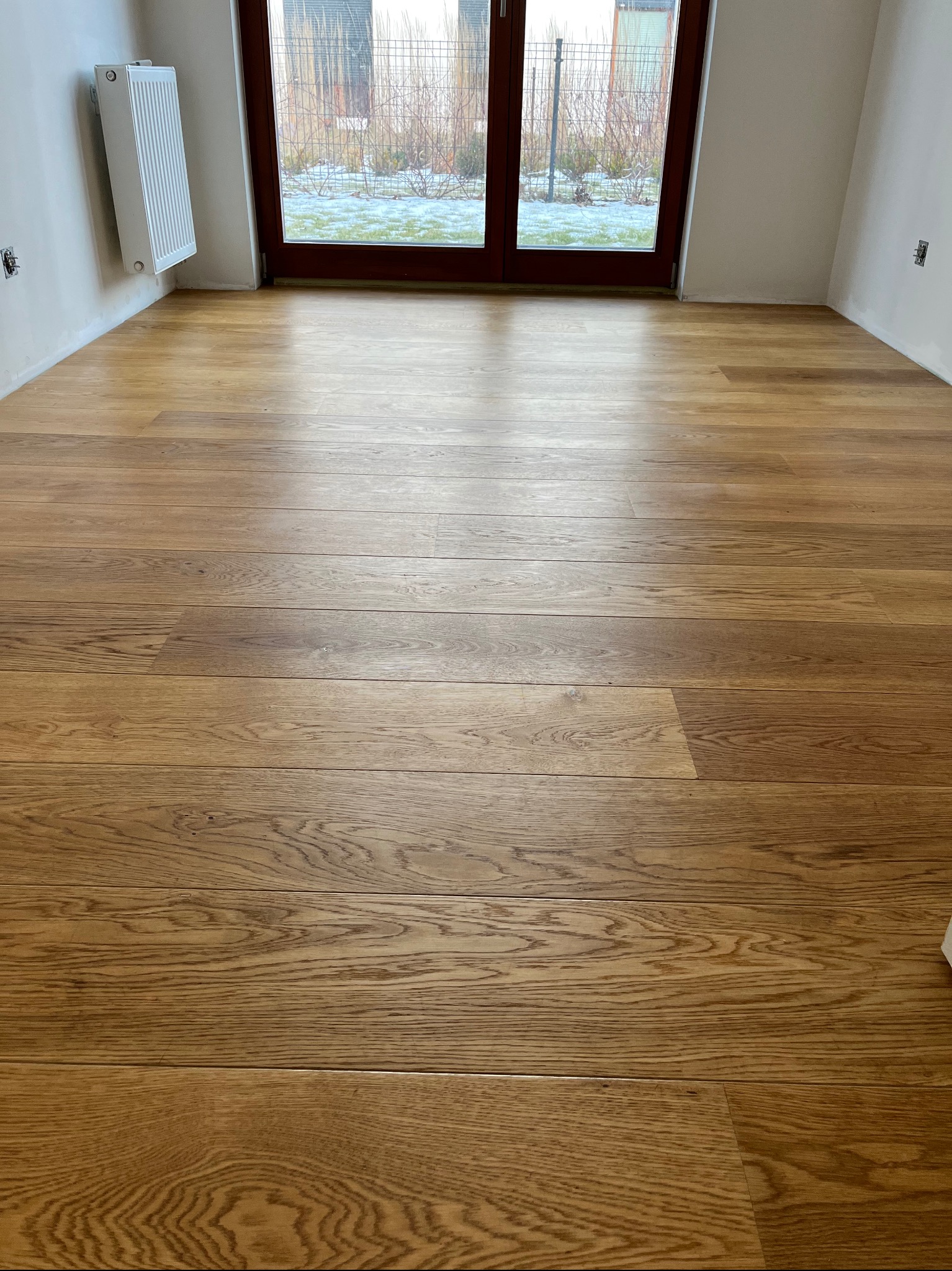 Jasna, dębowa podłoga parkietowa ułożona w nowym pomieszczeniu z grzejnikiem i dużym oknem balkonowym z widokiem na ogród zimą.