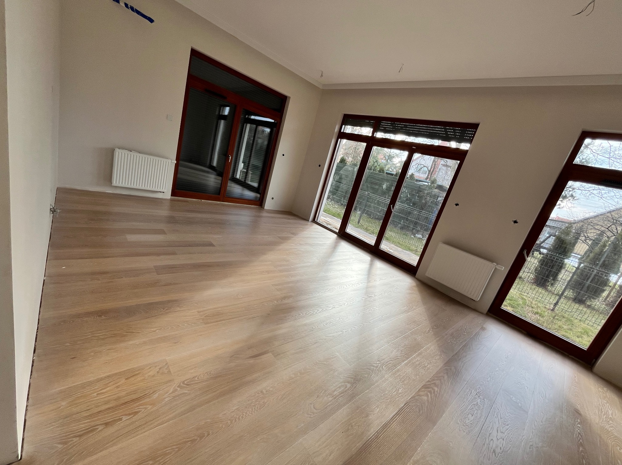 Jasny, świeżo położony parkiet w przestronnym pokoju z dużymi oknami w ciemnych ramach. Widoczne grzejniki pod oknami i surowe ściany, sugerujące stan wykończeniowy.