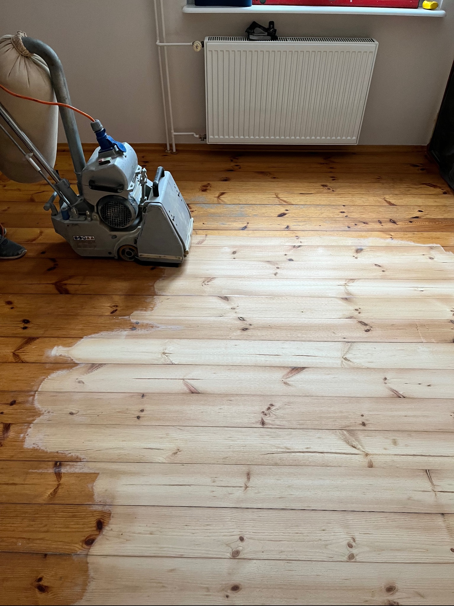 Parkiet z sosnowych desek częściowo wyszlifowany maszyną do cyklinowania, widoczny kontrast między surowym drewnem a wykończoną powierzchnią, w tle grzejnik.