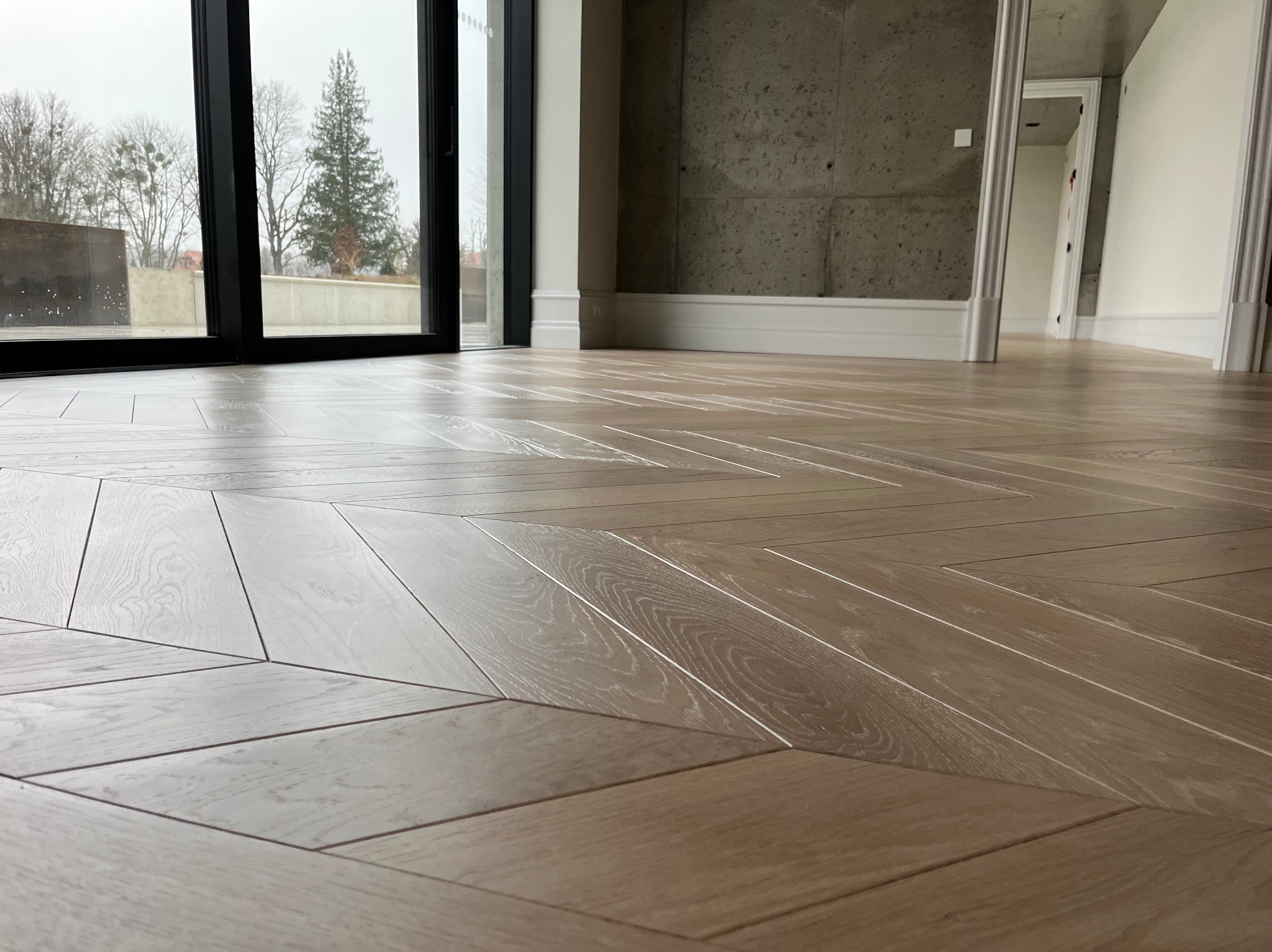 Jasny parkiet ułożony we wzór jodełki francuskiej w nowoczesnym wnętrzu z dużym oknem i betonową ścianą.