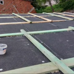 Czarna papa na dachu z drewnianym rusztem, widoczne buty robotnika i plastikowe wiaderko z elementami mocującymi