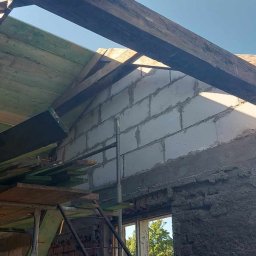 Adowicz remonty - Surowy stan wnętrza w trakcie remontu: widoczne ściany z bloczków, fragmenty starego muru pokrytego zaprawą, drewniana konstrukcja dachu i rusztowanie.