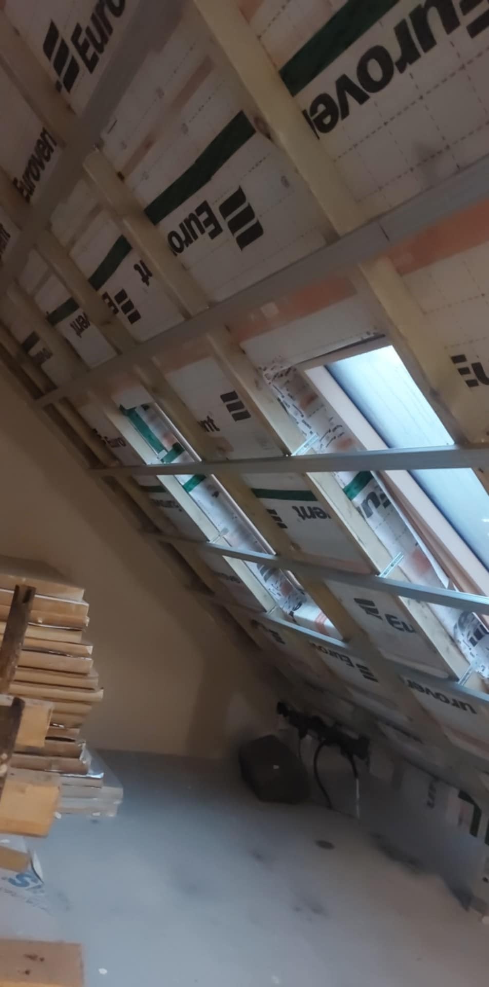 Montaż izolacji termicznej Euroven na poddaszu z widoczną konstrukcją drewnianą i oknem dachowym, w tle stos desek.