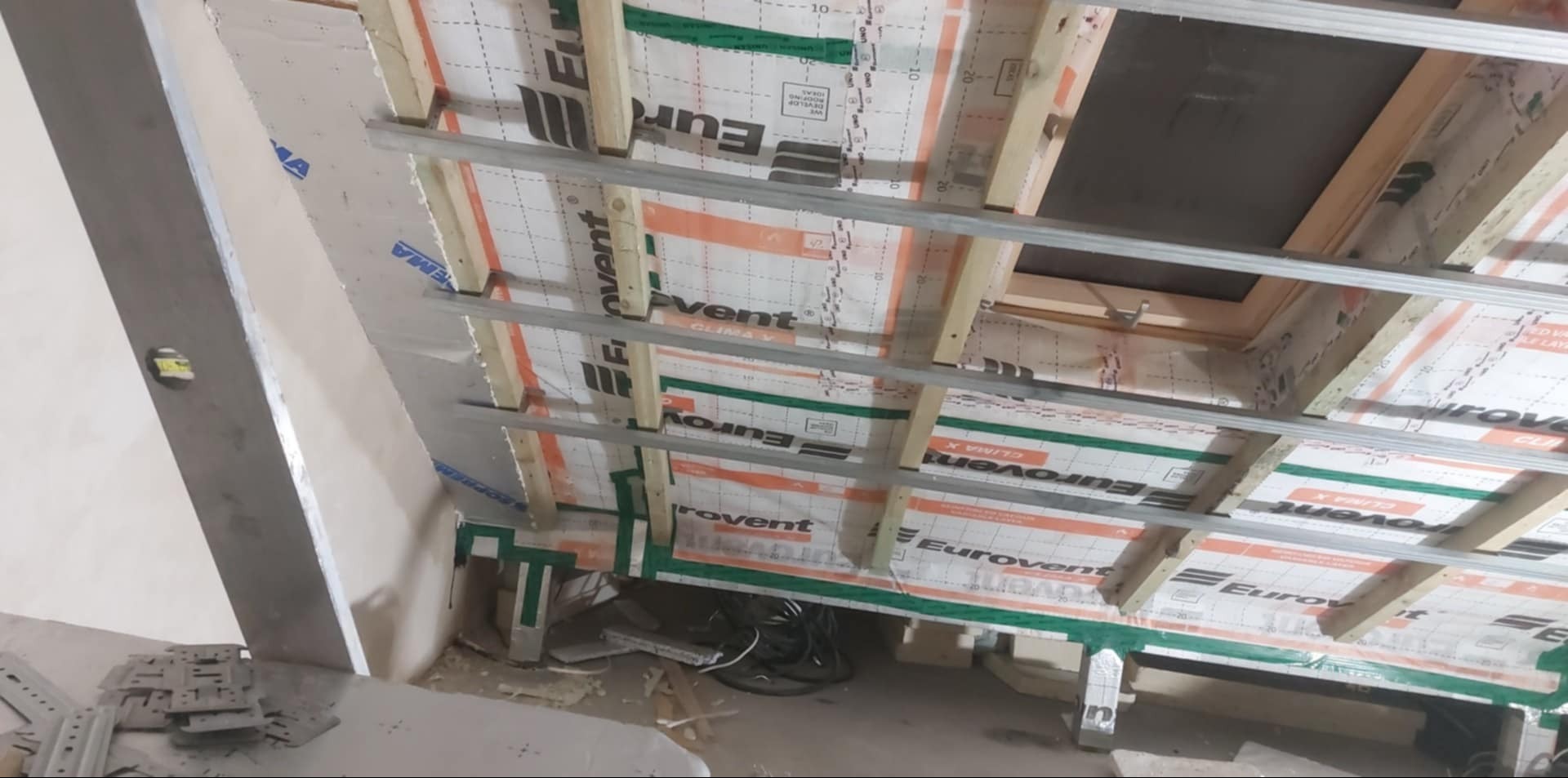 Sufit poddasza w trakcie izolacji, widoczne drewniane belki, profile metalowe i folia paroizolacyjna Eurovent Clima X. Przewody elektryczne leżące na podłodze.