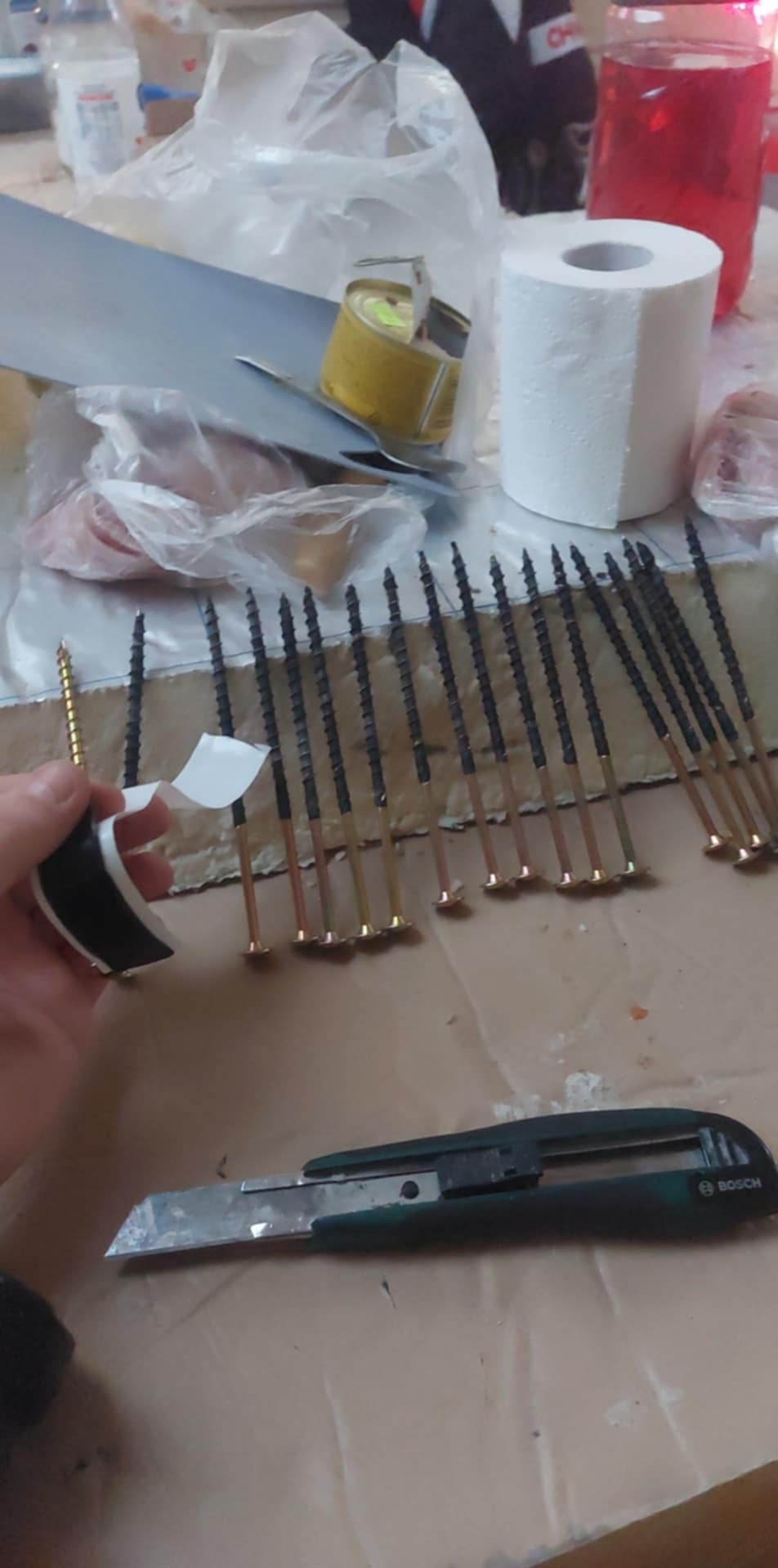 Przygotowanie do montażu: rząd śrub z czarną taśmą izolacyjną, obok nóż monterski Bosch, w tle rolka papieru toaletowego i napój w butelce.