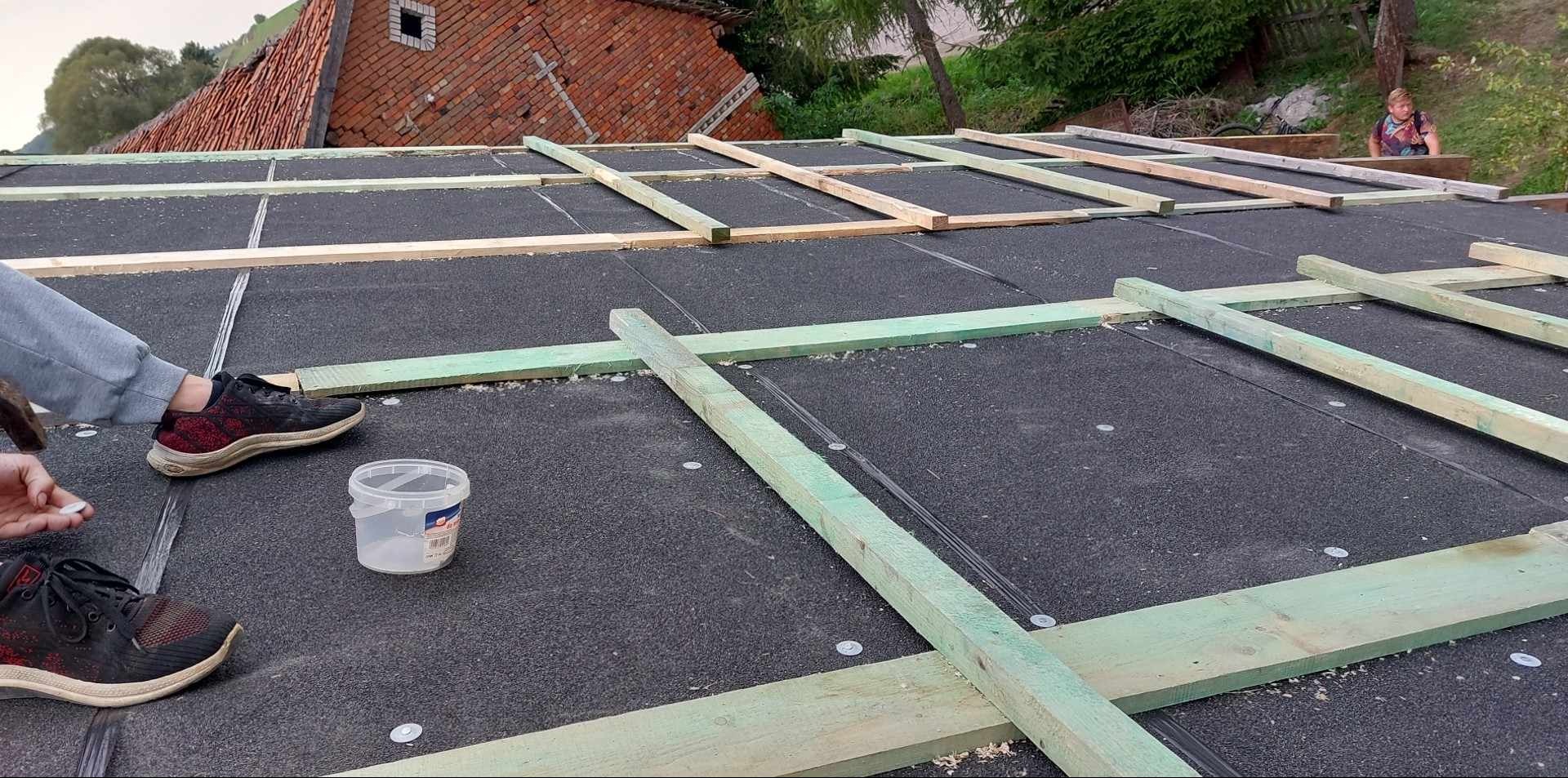 Czarna papa na dachu z drewnianym rusztem, widoczne buty robotnika i plastikowe wiaderko z elementami mocującymi