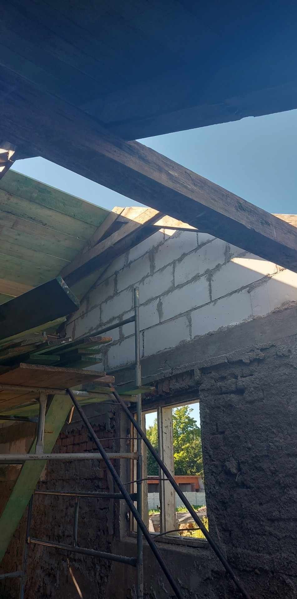 Surowy stan wnętrza w trakcie remontu: widoczne ściany z bloczków, fragmenty starego muru pokrytego zaprawą, drewniana konstrukcja dachu i rusztowanie.