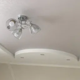 Wykończony sufit podwieszany z oświetleniem punktowym i ozdobną lampą z metalowymi kloszami, widoczne ślady szpachlowania na ścianach.