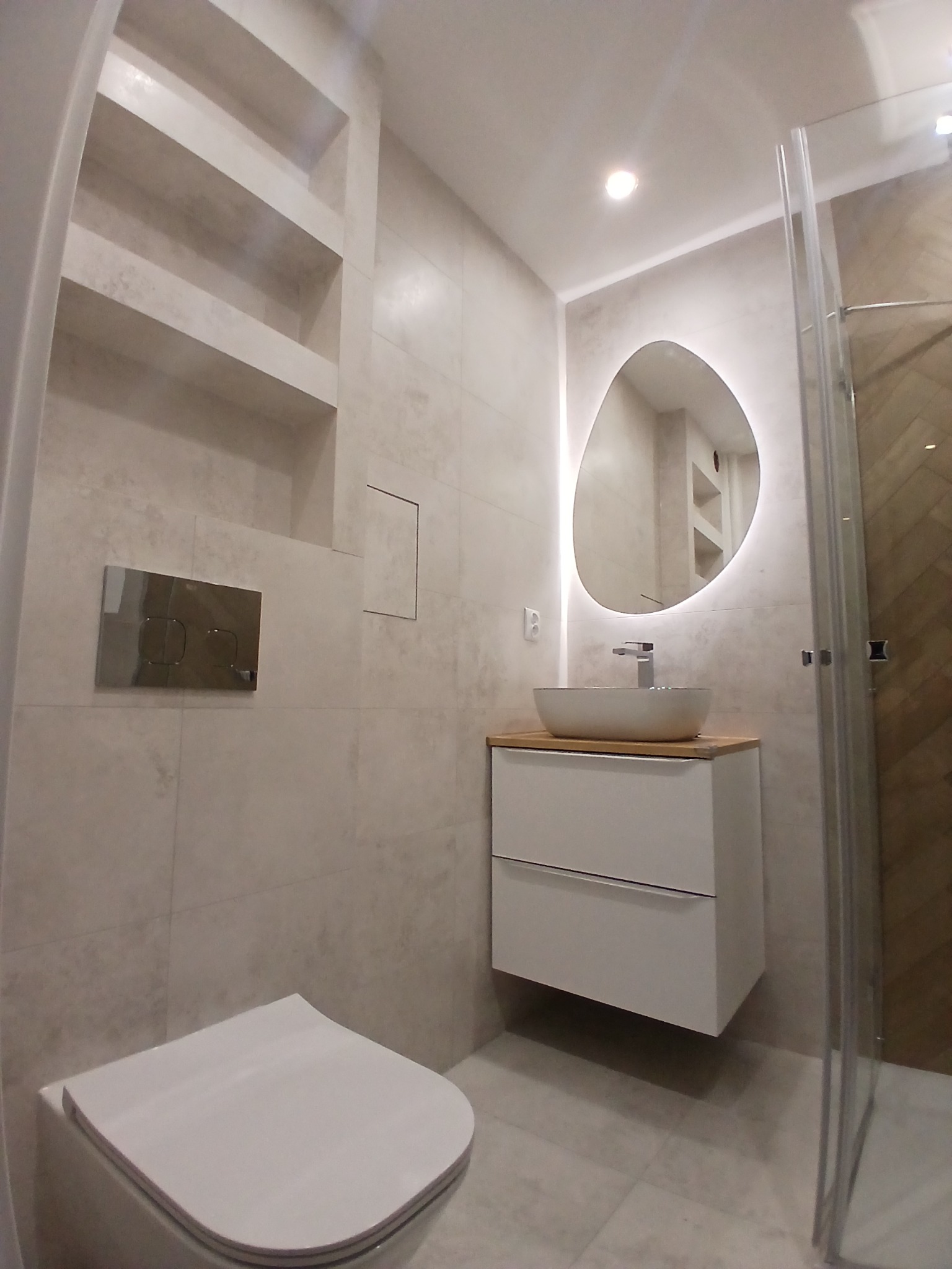 Nowoczesna łazienka po remoncie: beżowe płytki, podświetlane lustro o nieregularnym kształcie, wisząca szafka z umywalką i kabina prysznicowa z elementami drewna.