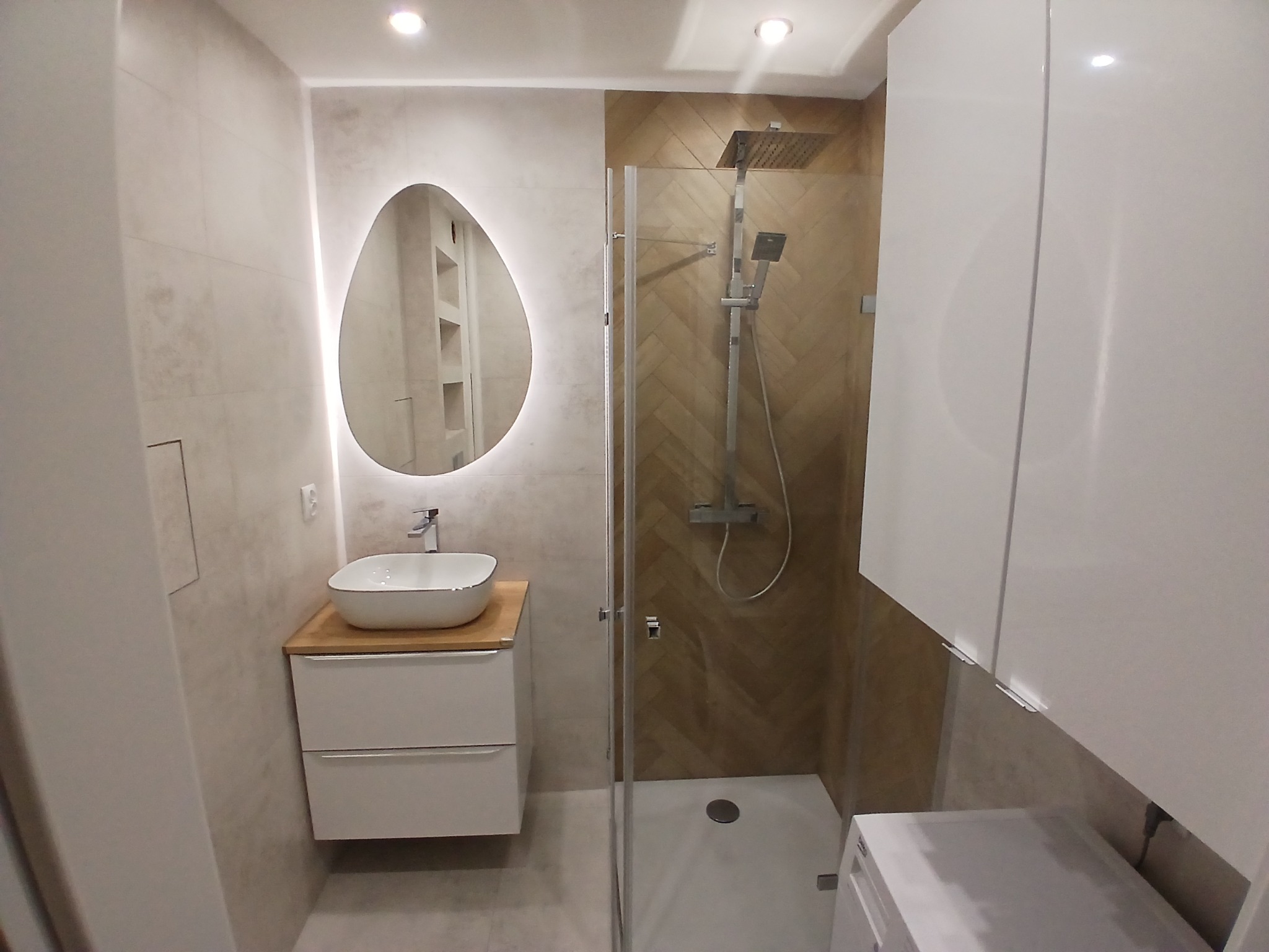 Nowoczesna łazienka z owalnym lustrem podświetlanym LED, kabiną prysznicową typu walk-in i białą szafką z umywalką nablatową. Płytki w odcieniach beżu i brązu.