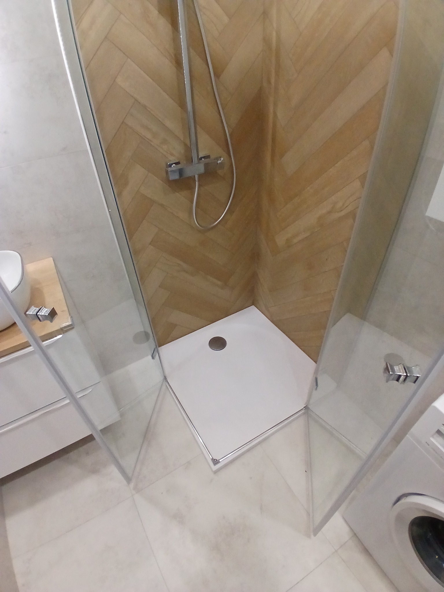 Nowoczesna kabina prysznicowa z brodzikiem i armaturą, wykończona płytkami imitującymi drewno ułożonymi w jodełkę. Widoczna umywalka i pralka.