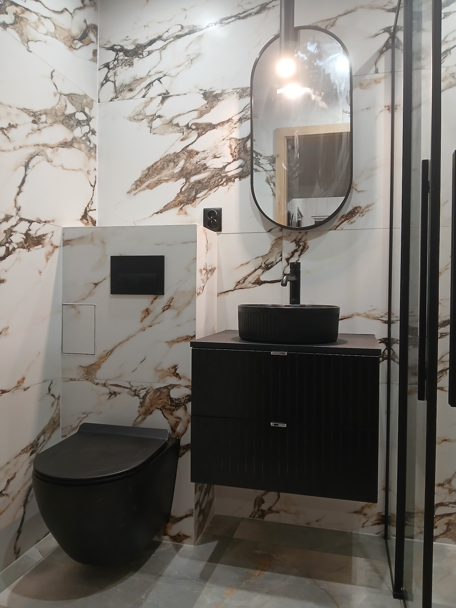 Wykończona łazienka w stylu nowoczesnym: czarna armatura, umywalka nablatowa, wisząca toaleta i płytki imitujące marmur z brązowymi żyłami.
