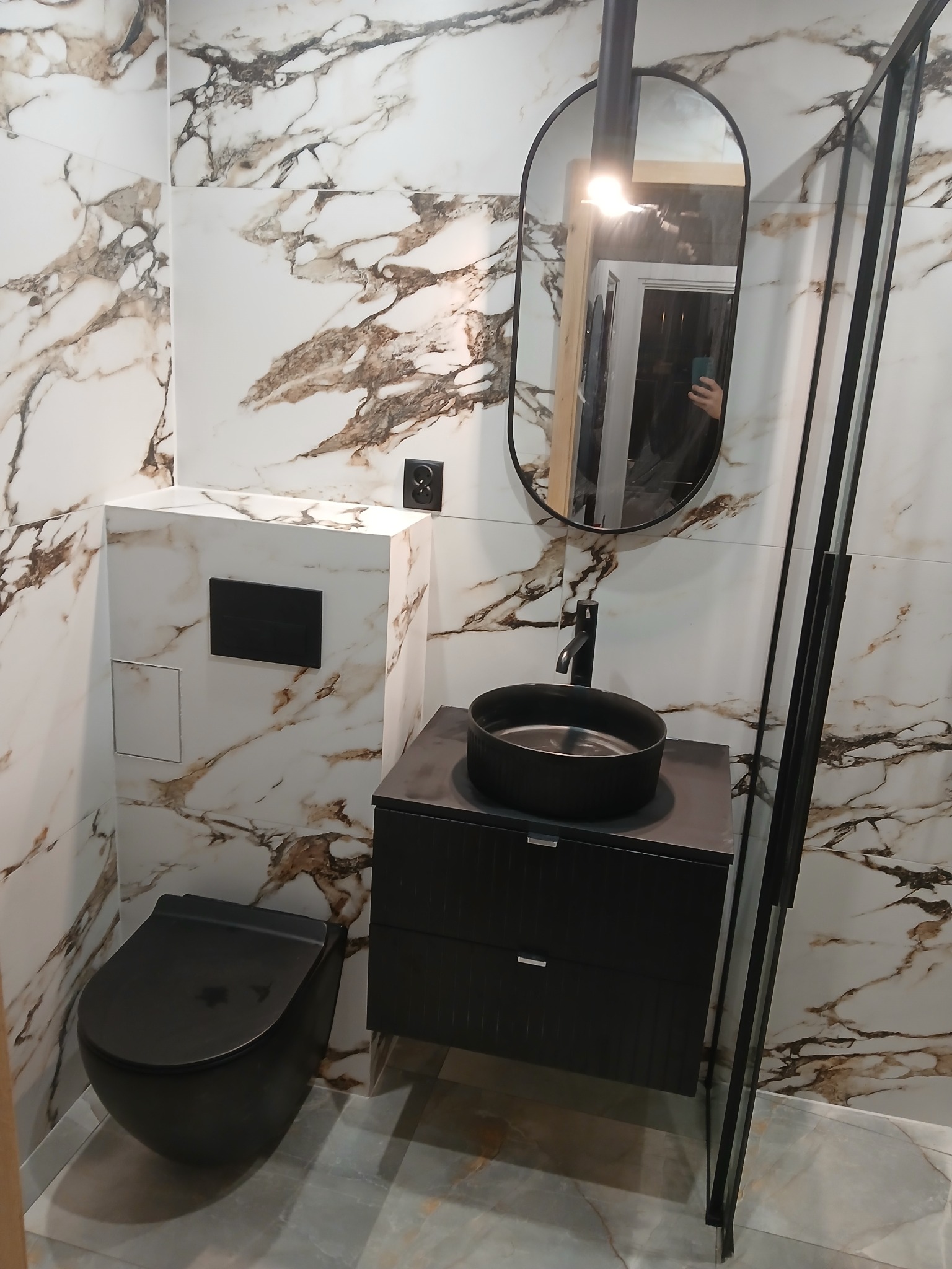 Nowoczesna łazienka z czarną armaturą: umywalka nablatowa, toaleta podwieszana. Ściany wykończone płytkami imitującymi marmur. Minimalistyczne lustro.