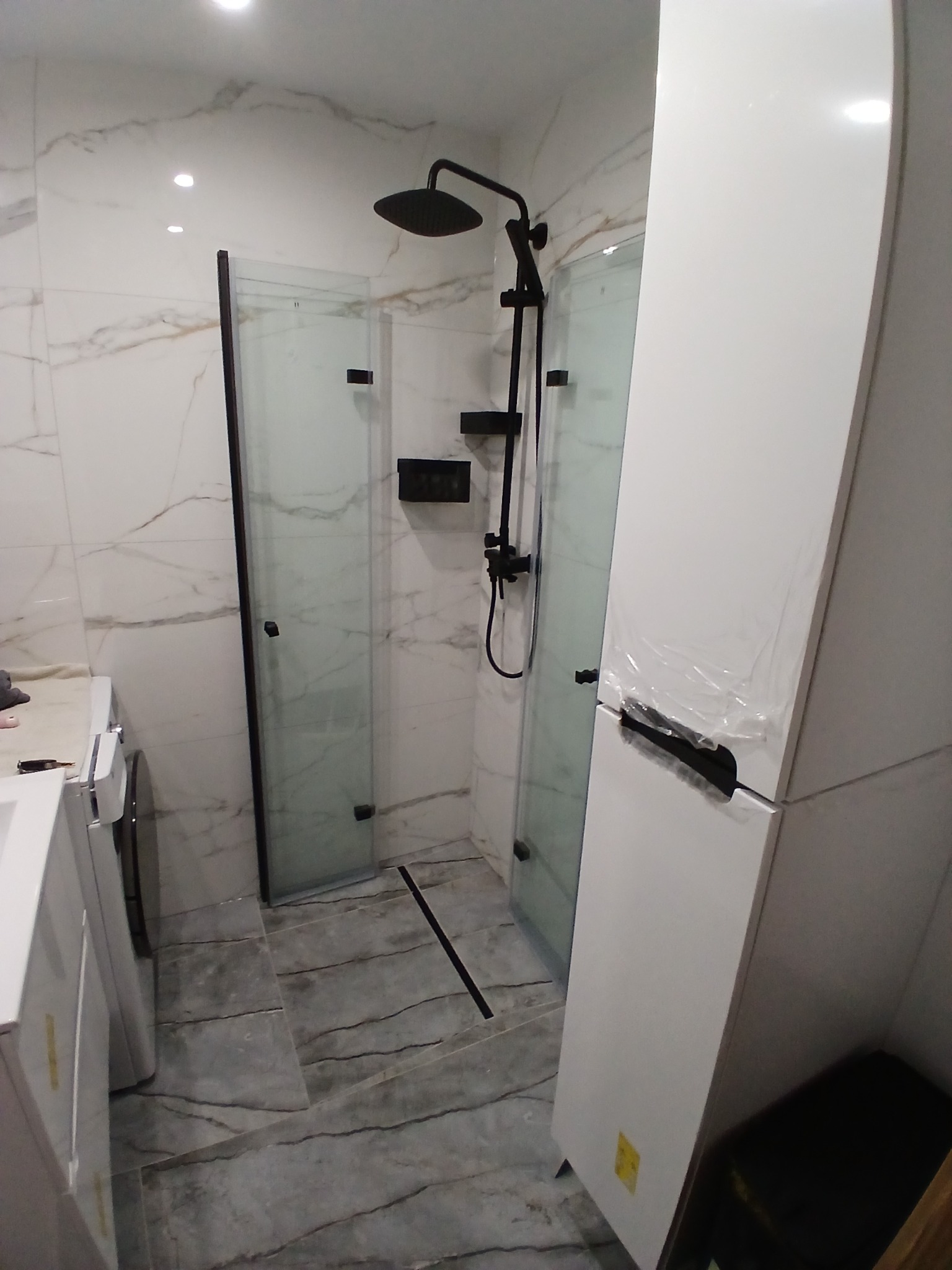 Nowoczesna kabina prysznicowa z czarną armaturą i szklanymi drzwiami w łazience wykończonej marmurowymi płytkami. Widoczna biała szafka łazienkowa.