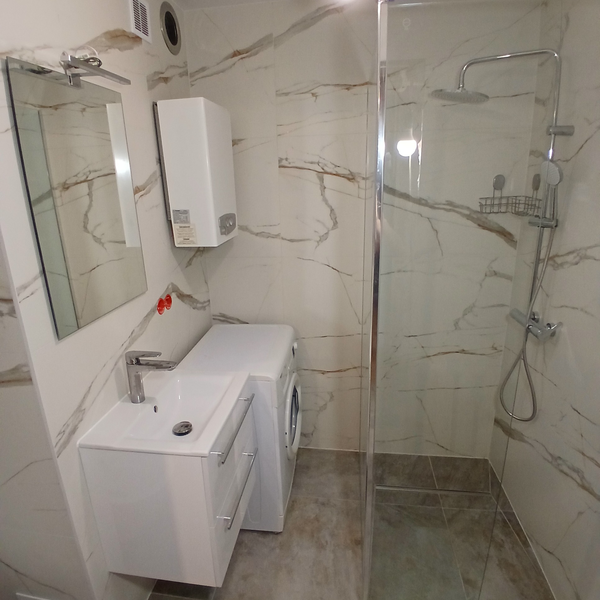 Nowoczesna łazienka z kabiną prysznicową typu walk-in, umywalką z szafką i pralką. Marmurowe płytki na ścianach i podłodze. Minimalistyczny design.