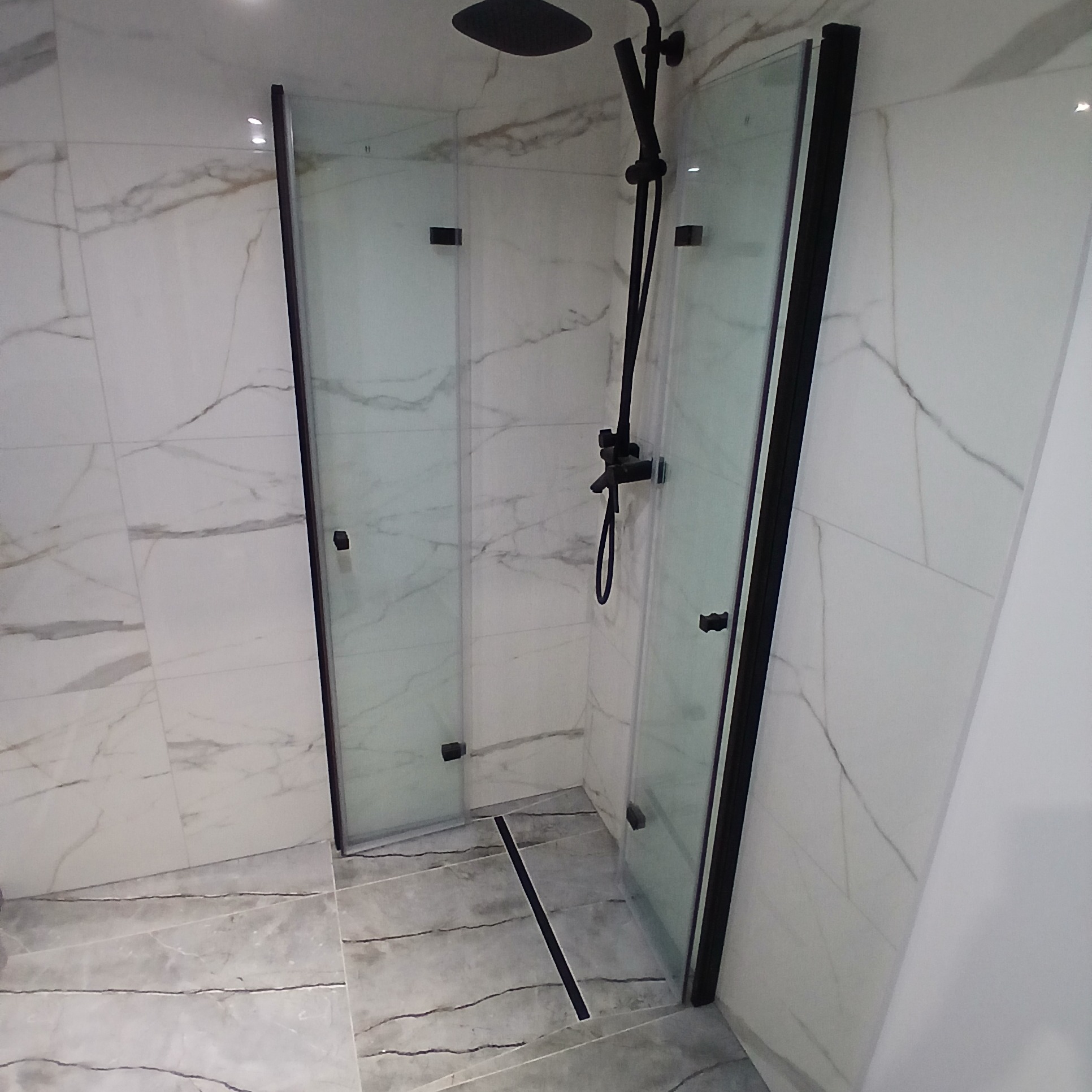 Nowoczesna kabina prysznicowa z czarnymi profilami i deszczownicą, wykończona marmurowymi płytkami. Minimalistyczny design z odpływem liniowym.