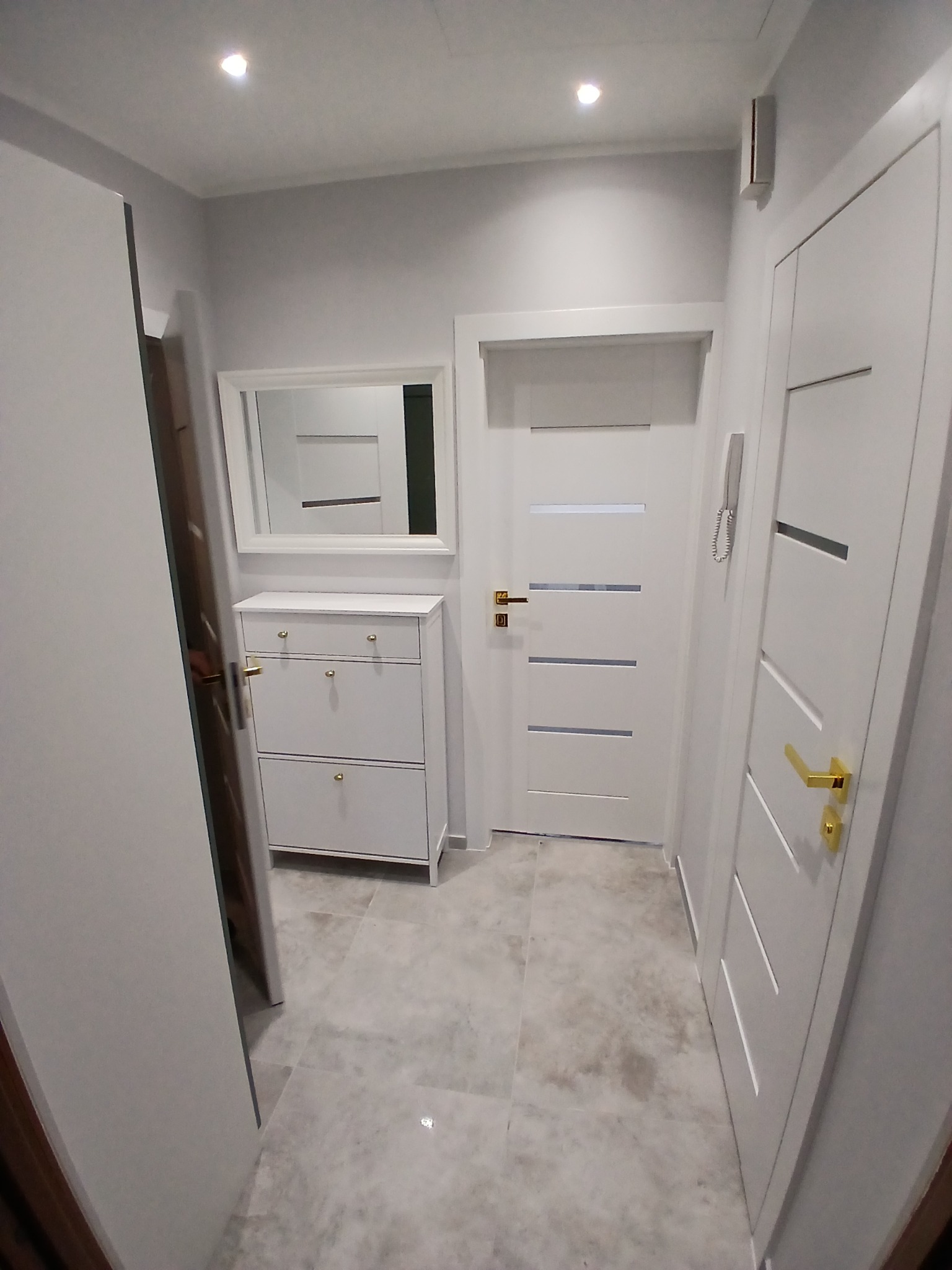 Jasne wnętrze przedpokoju z białymi drzwiami z poziomymi przeszkleniami i złotymi klamkami, szafką na buty z lustrem i szarą podłogą z płytek.