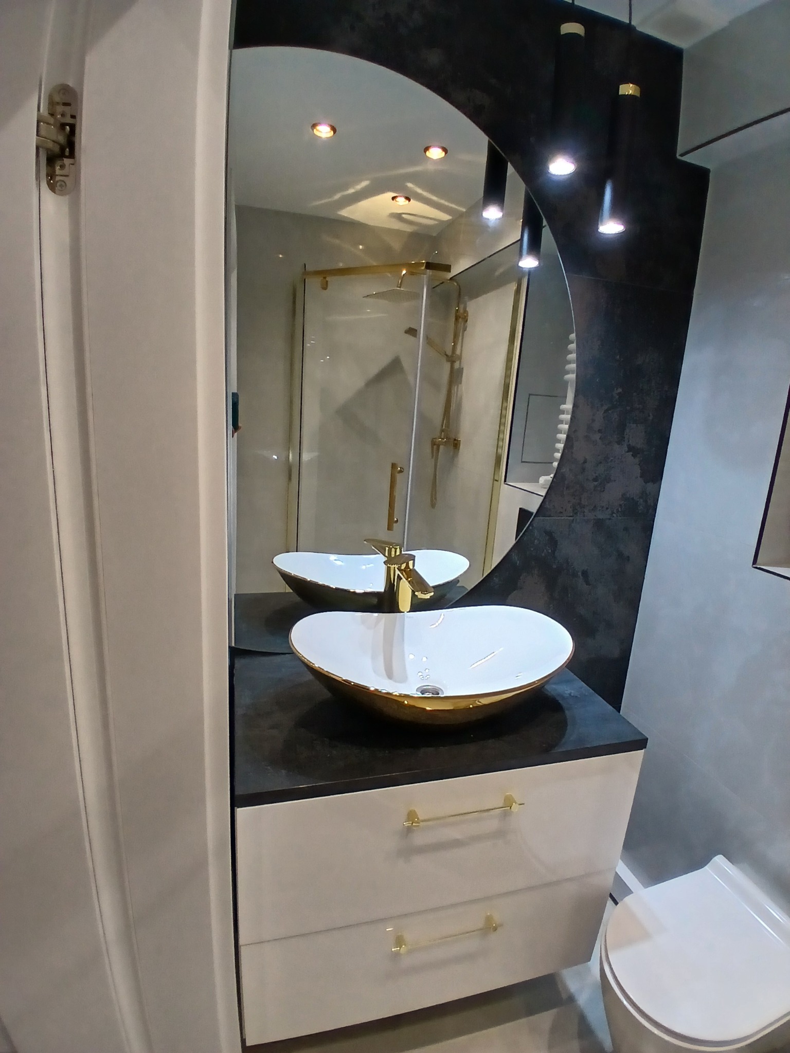 Nowoczesna łazienka z owalną umywalką w kolorze białym ze złotymi akcentami, czarnym blatem i szafką z białymi frontami. W tle kabina prysznicowa i lustro w kształcie łuku.