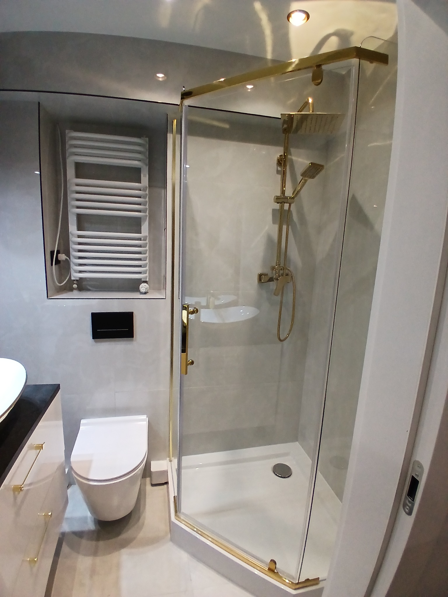Nowoczesna łazienka z kabiną prysznicową w złotych ramach, wiszącą toaletą i grzejnikiem drabinkowym w niszy. Minimalistyczny design, szare płytki i złote akcenty.