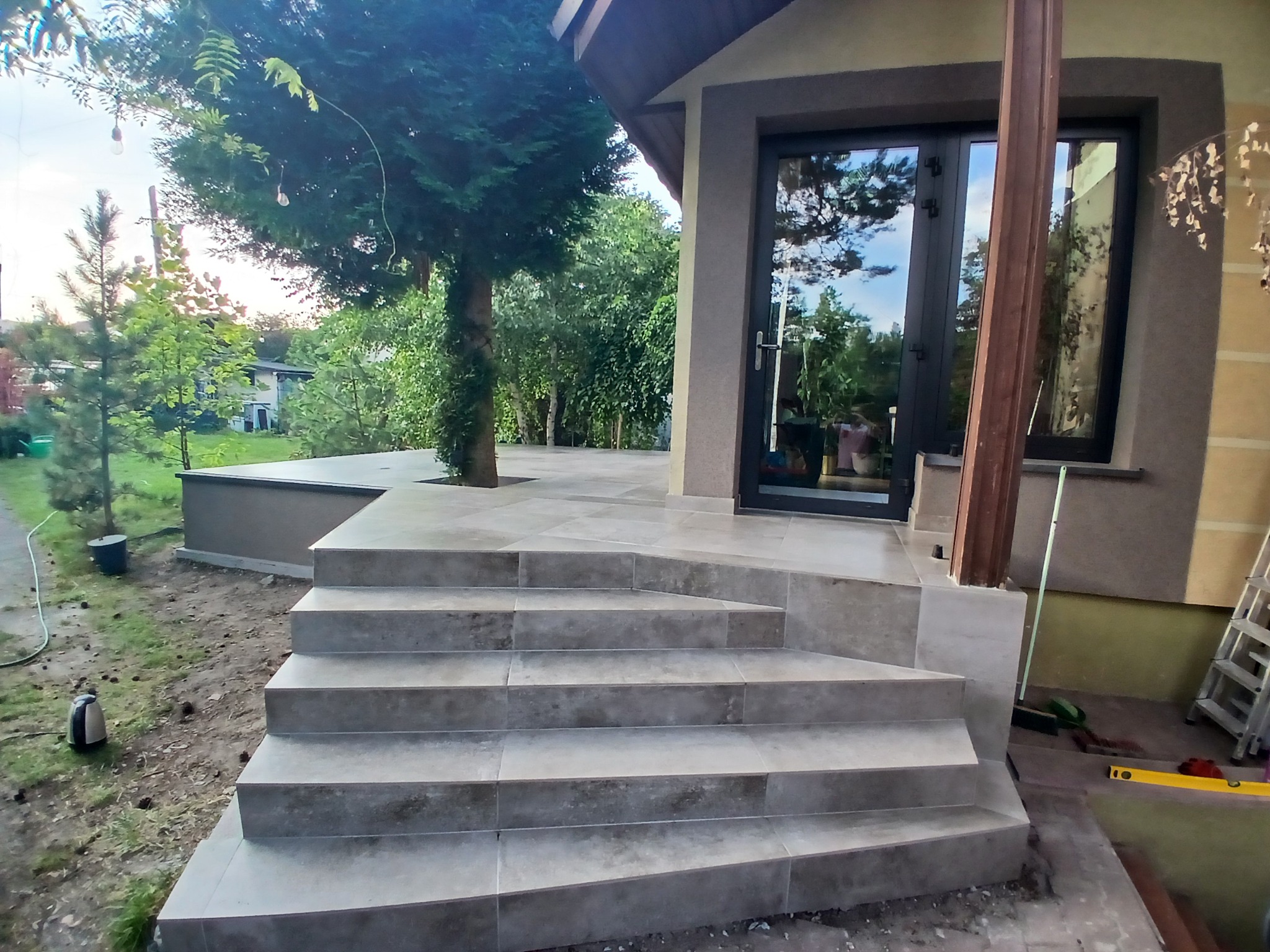 Wykończone schody zewnętrzne z szarych płytek prowadzące do domu z ciemnymi drzwiami. Widoczne narzędzia i zieleń w tle. Ujęcie z dołu, podkreślające solidność wykonania.