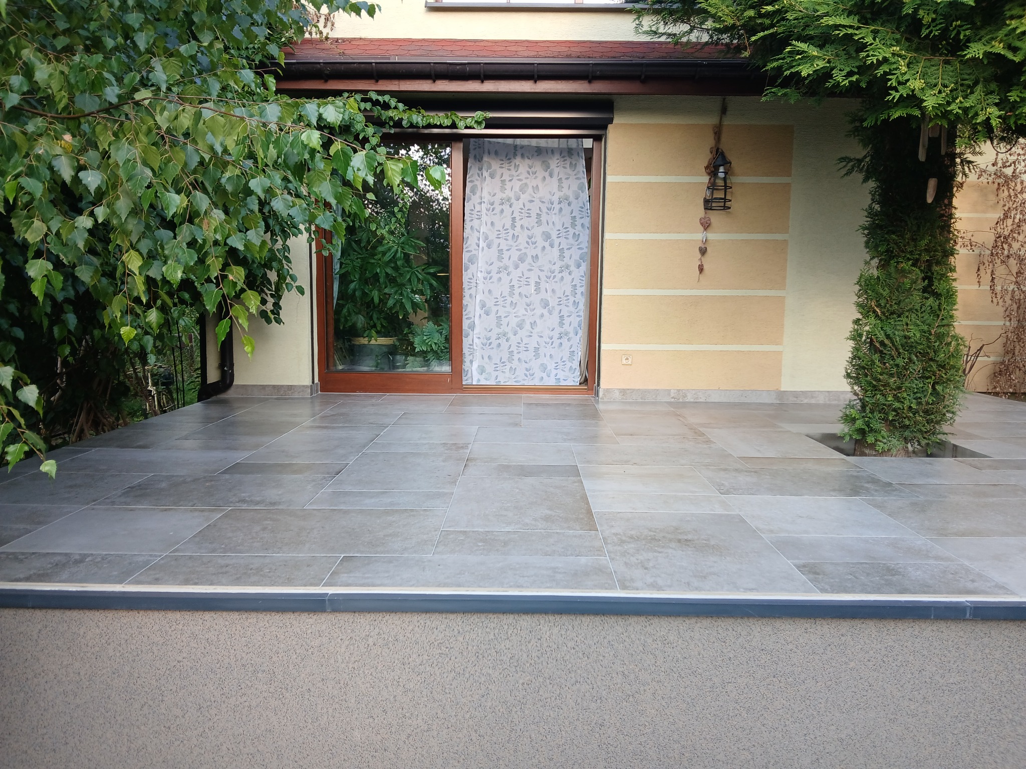 Wyłożony płytkami taras w Katowicach z widokiem na zieleń. Płytki w odcieniach szarości, ułożone w prostokątnym wzorze. Drewniane drzwi tarasowe z zasłoną w tle.