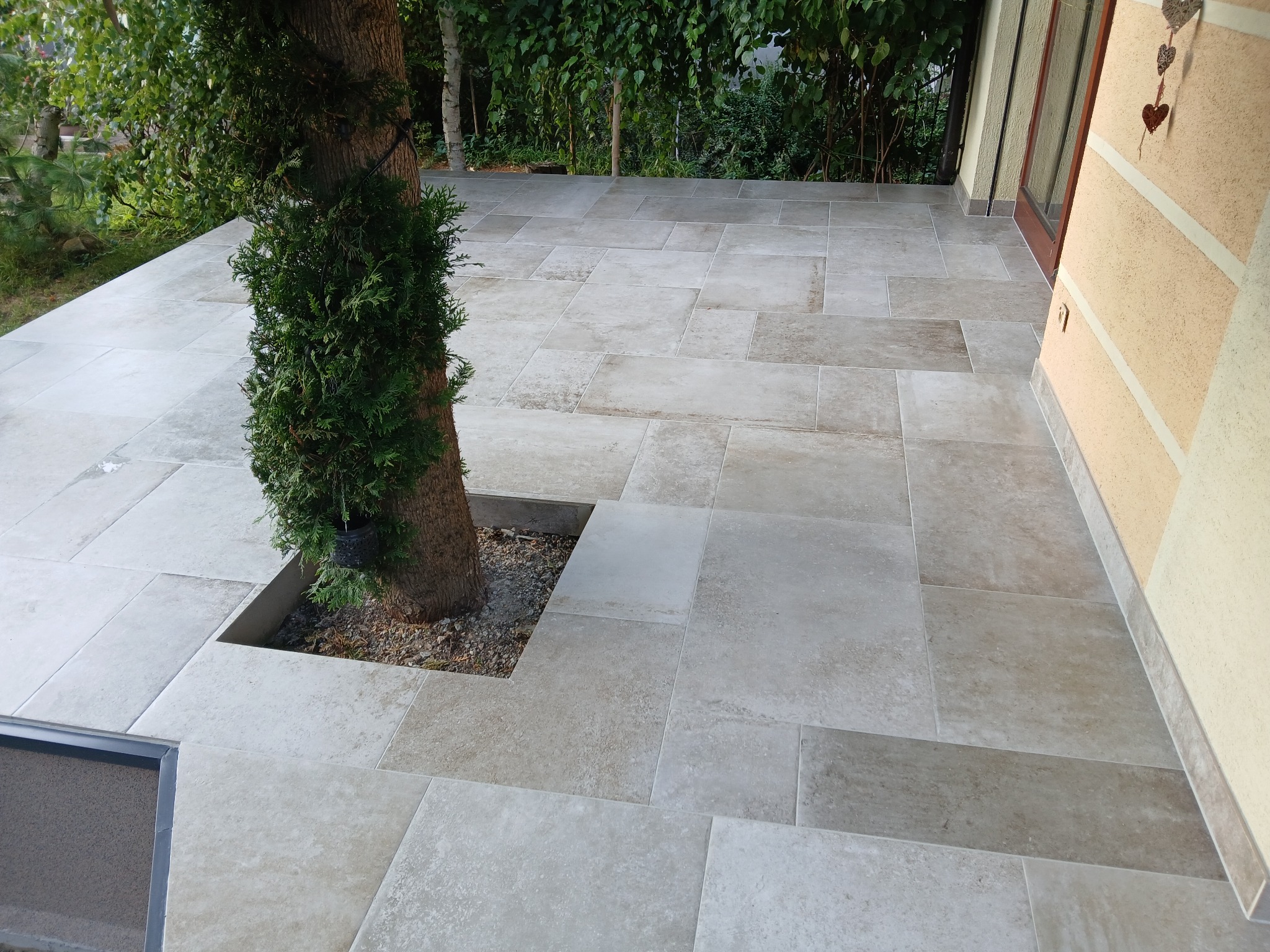 Jasne, nieregularne płytki tarasowe ułożone wokół drzewa, z widocznym fragmentem elewacji budynku i zielenią w tle. Ujęcie z góry, podkreślające wzór i fakturę płytek.