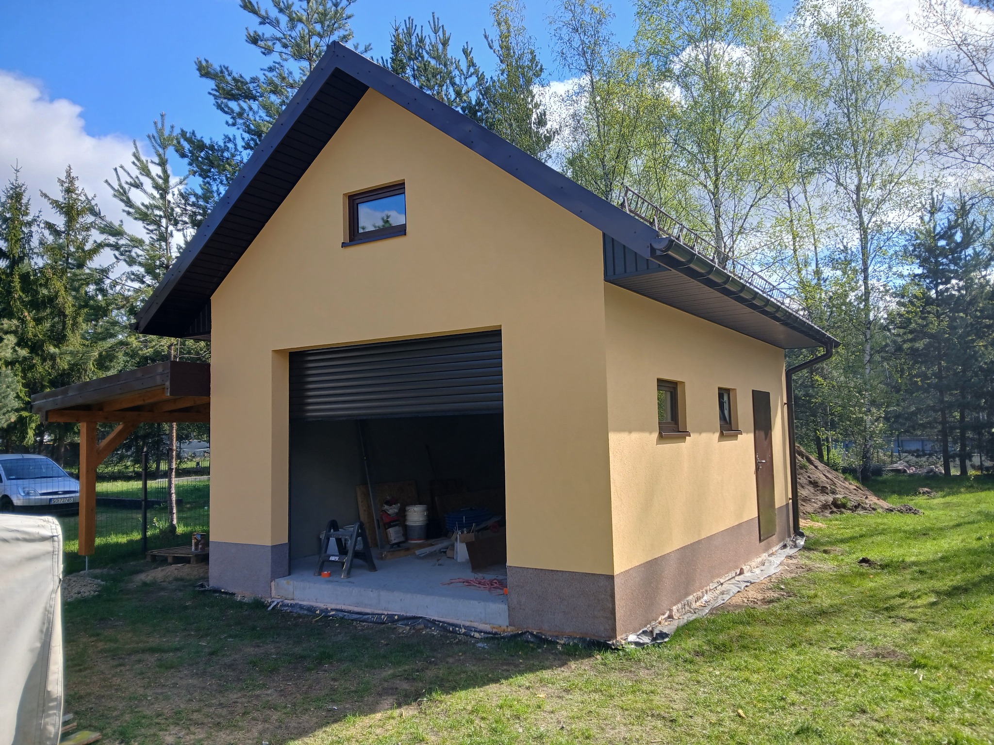 Otynkowany garaż wolnostojący z brązowym cokołem i czarnym dachem na tle zieleni. Widoczne okna, brama garażowa i drzwi wejściowe. Słoneczny dzień.