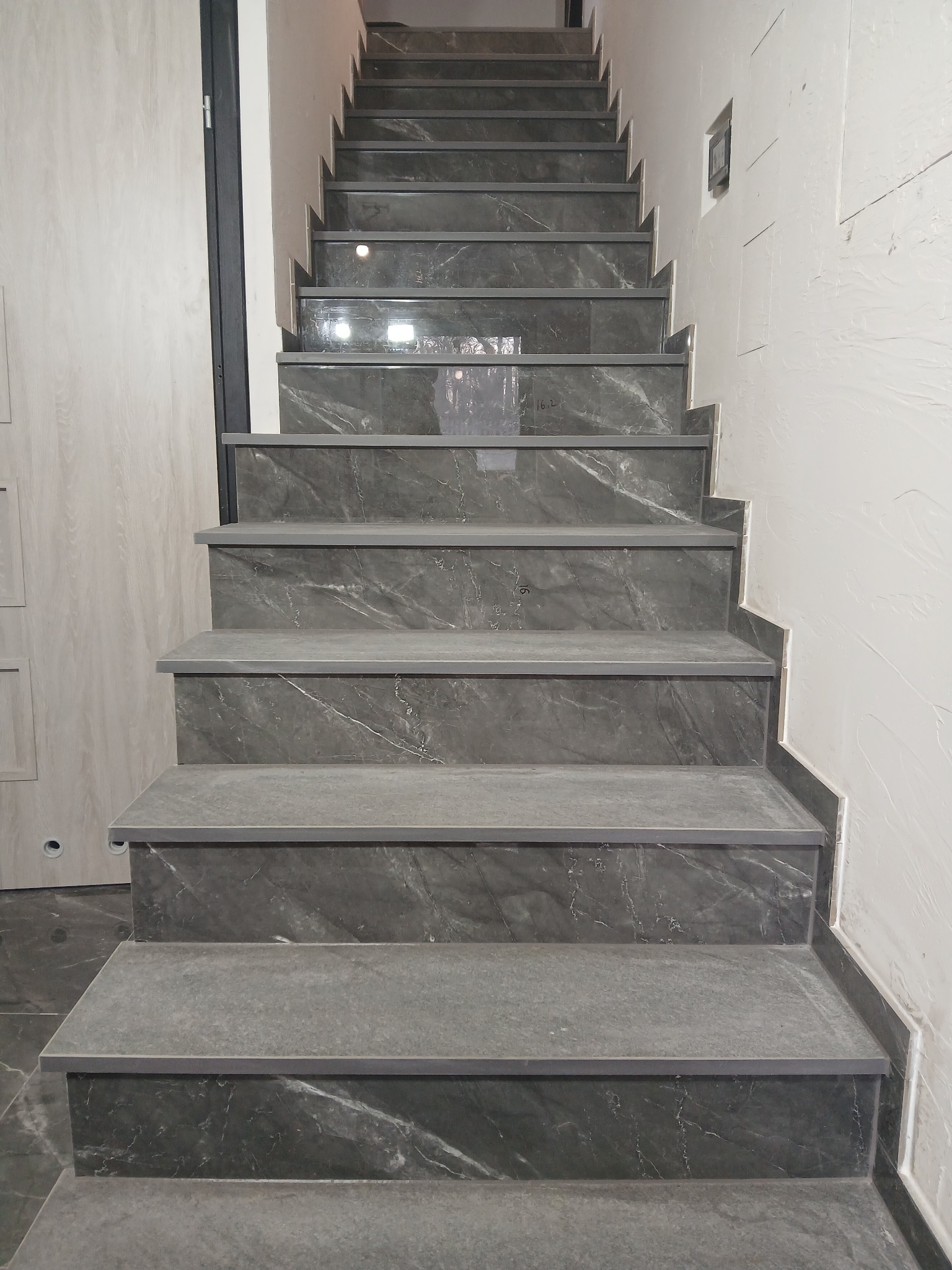 Schody wykończone szarą płytką imitującą marmur, widok z dołu. Widoczne niedoskonałości na ścianie wymagające wykończenia. Nowoczesny design.