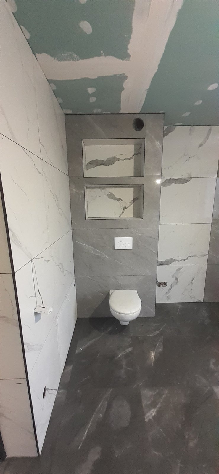 Wykończona łazienka z szarymi i białymi płytkami imitującymi marmur, podwieszana toaleta i wnęki w ścianie. Sufit z płyt karton-gips. Widok z dołu.