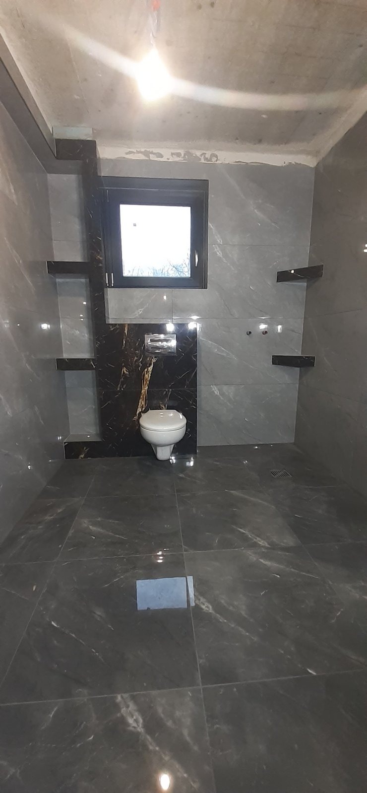 Wykończona łazienka z szarymi płytkami imitującymi marmur, czarnymi akcentami i podwieszaną toaletą. Widoczne półki i okno. Nowoczesny design.