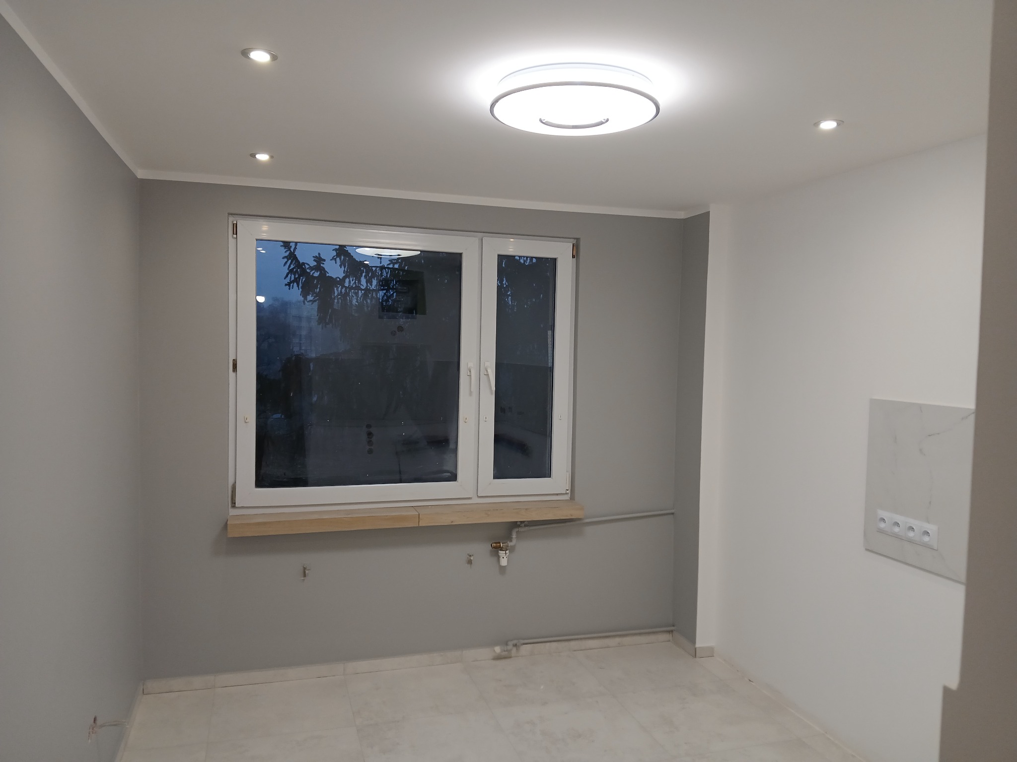 Jasne wnętrze po remoncie: szare ściany, białe okno z drewnianym parapetem, marmurowe płytki podłogowe, nowoczesne oświetlenie sufitowe i gniazdka elektryczne.