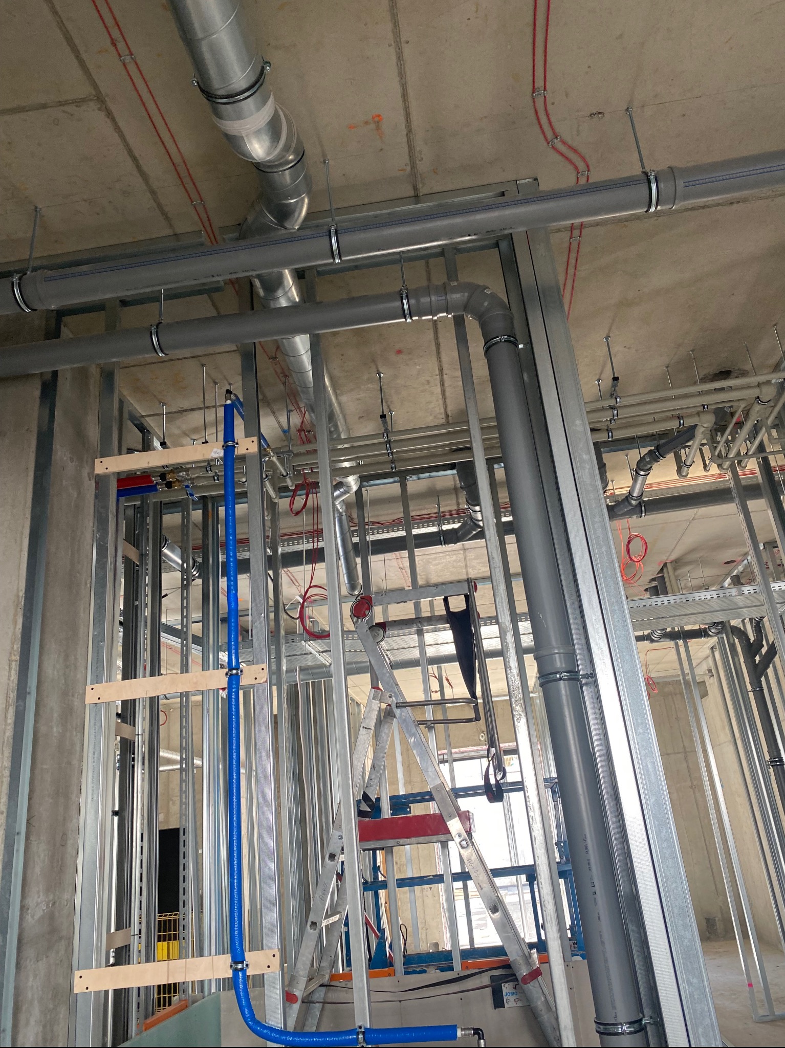 Instalacja wentylacyjna i hydrauliczna w surowym stanie, widoczne rury wentylacyjne, rury hydrauliczne, metalowa konstrukcja, drabina i narzędzia.