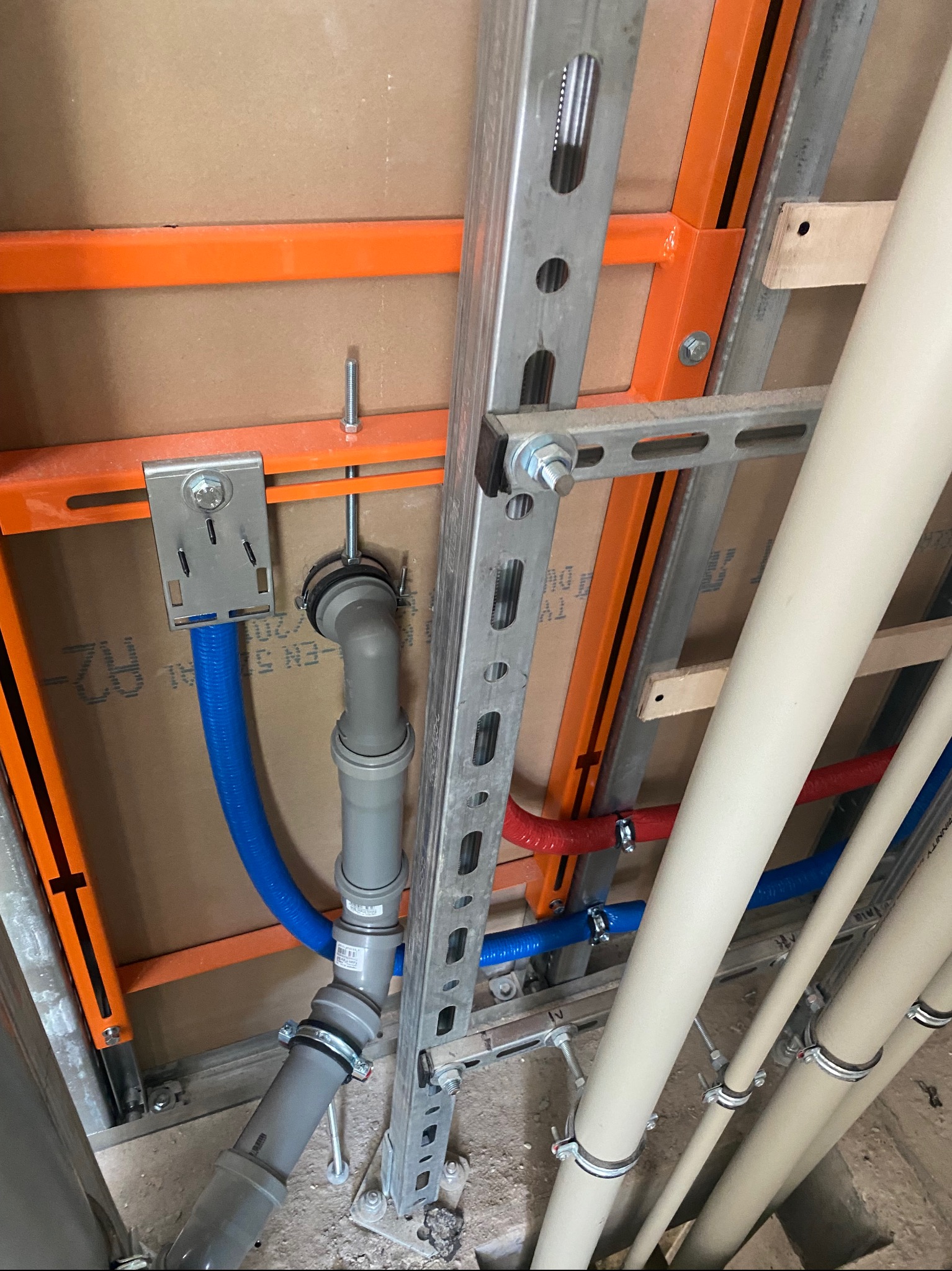 Szara rura kanalizacyjna z niebieską rurą wodną, zamontowane na pomarańczowym i srebrnym stelażu. Widoczne mocowania i elementy instalacji w surowym stanie.