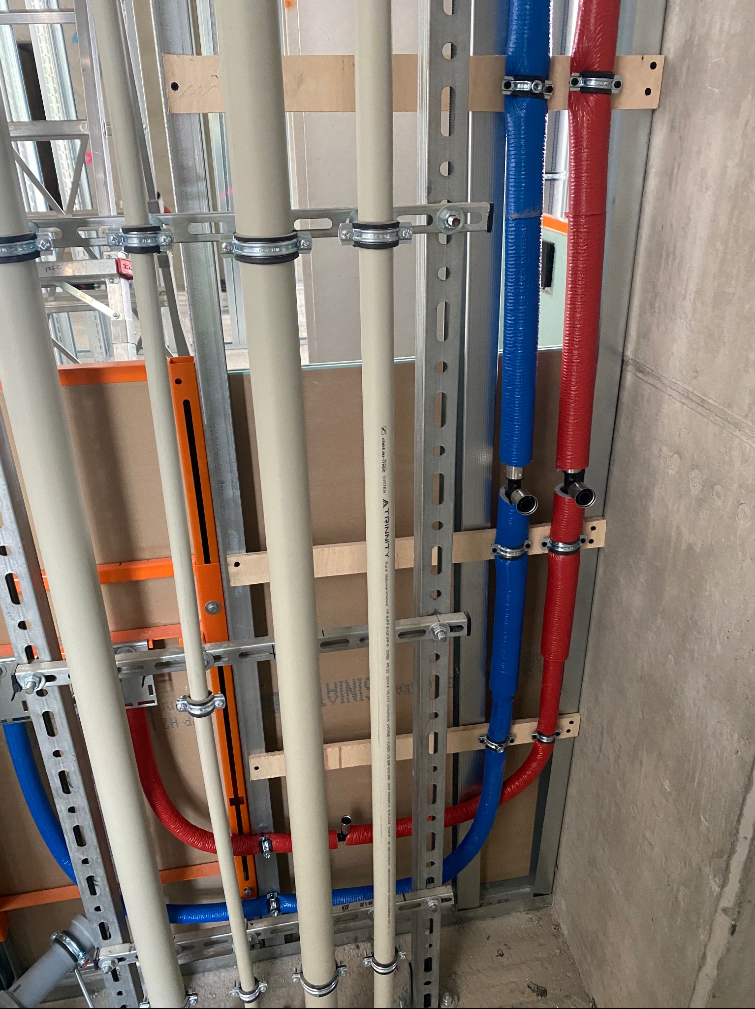 Instalacja rur wodnych i grzewczych w budynku w trakcie budowy. Widoczne rury w kolorach: beżowym, niebieskim i czerwonym, przymocowane do metalowej konstrukcji.
