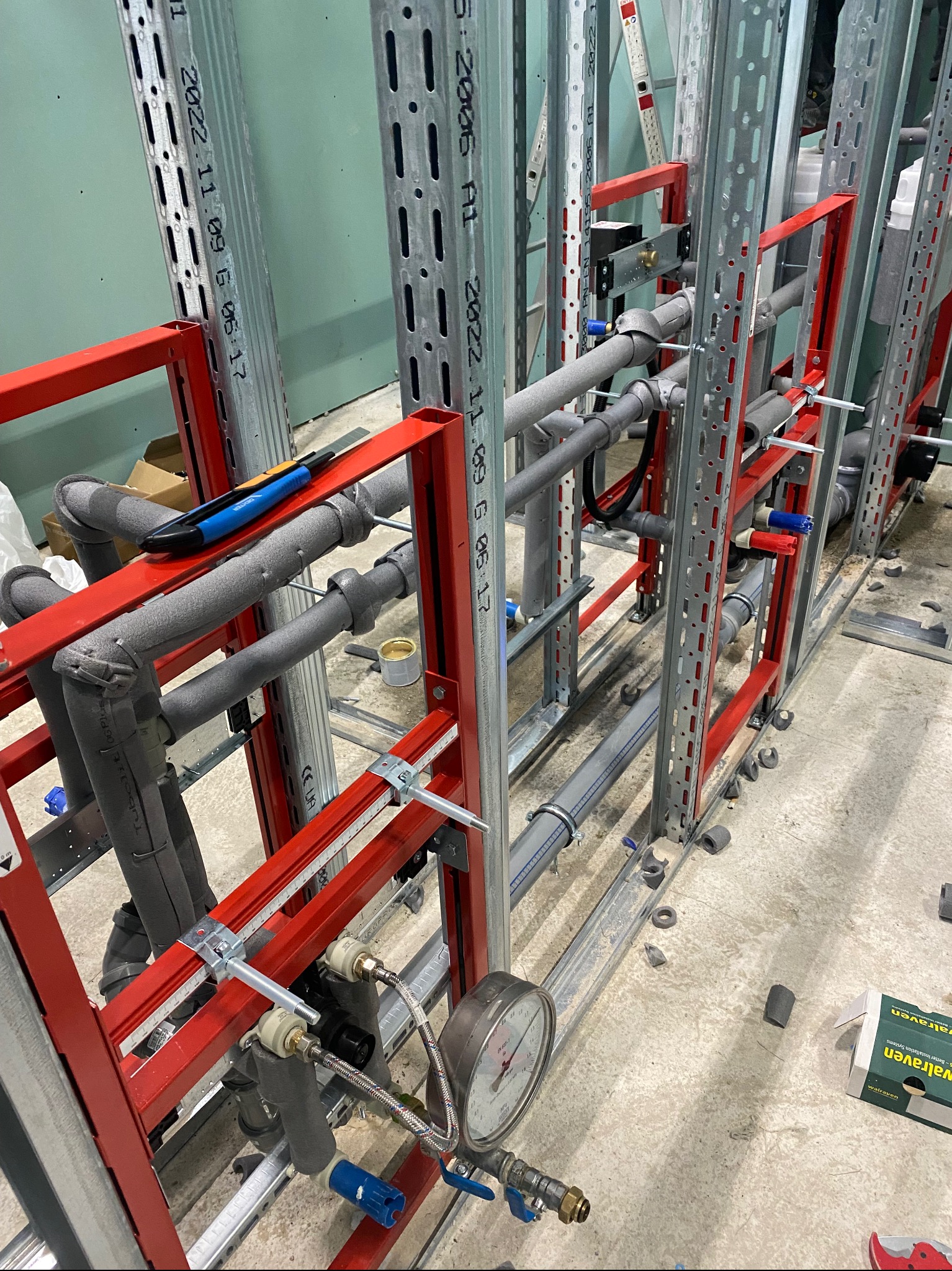 Montaż instalacji wodnej: rury w izolacji, manometr i metalowa konstrukcja wsporcza. Widoczne narzędzie i elementy montażowe na surowym podłożu.