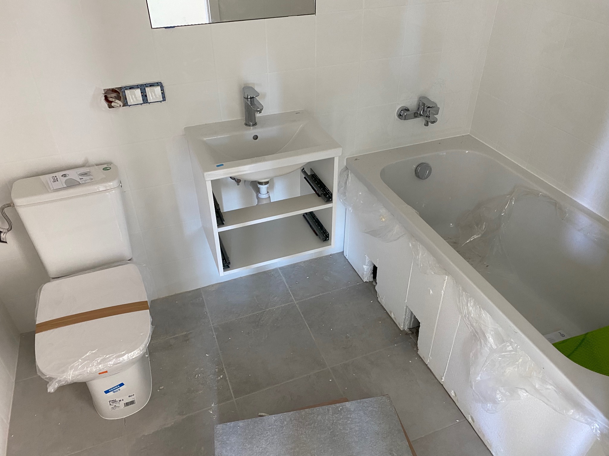 Nowa, jasna łazienka w trakcie wykańczania: biała armatura (umywalka z szafką, toaleta, wanna) częściowo zabezpieczona folią, szare płytki podłogowe, widoczne gniazdka.