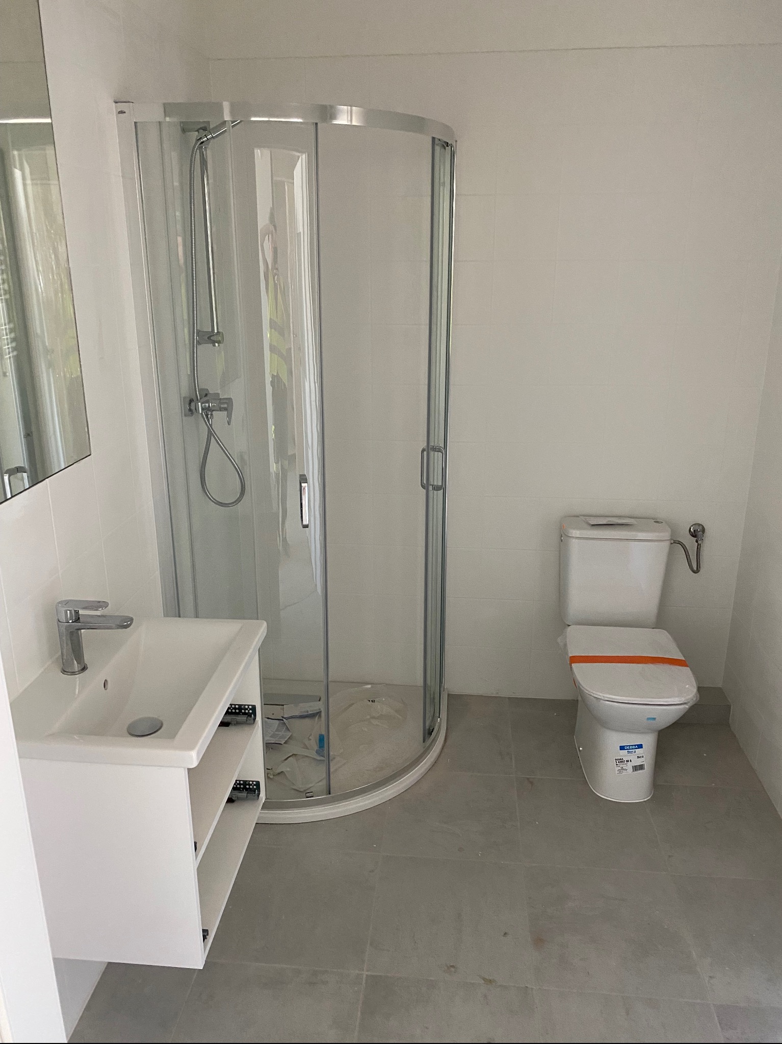 Nowoczesna, jasna łazienka po remoncie: kabina prysznicowa z chromowanymi detalami, biała umywalka z szafką i toaleta z pomarańczową taśmą zabezpieczającą.