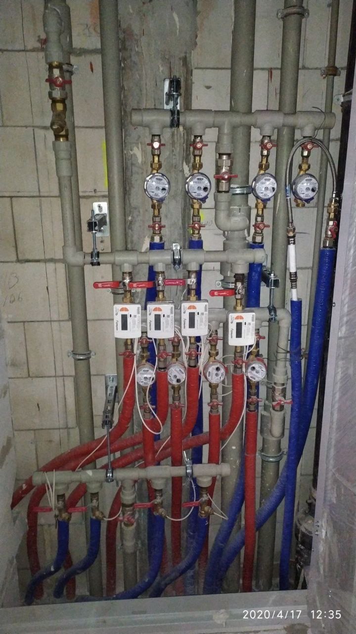 Rozbudowana instalacja wodna z licznymi rurami (czerwone i niebieskie) i zaworami, zamontowana na tle surowej ściany z płytek. Widoczne liczniki i elementy mocujące.