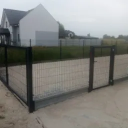 Nowoczesne, grafitowe ogrodzenie z siatki z furtką na betonowej podmur&oacute;wce, widok na dom w tle.