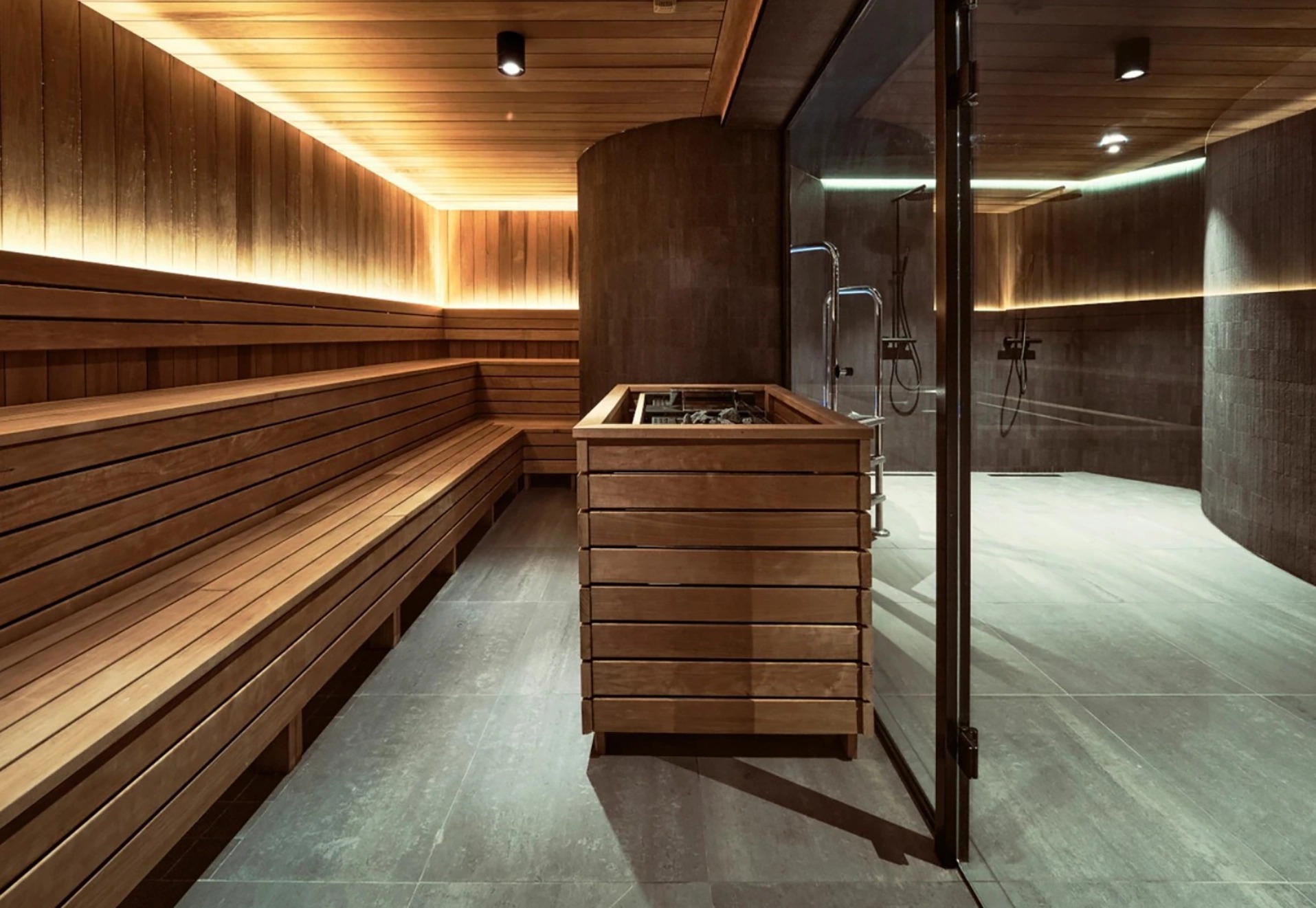 Nowoczesna sauna z drewnianymi ławkami, piecem obudowanym drewnem i przylegającą kabiną prysznicową oddzieloną szklanymi drzwiami.