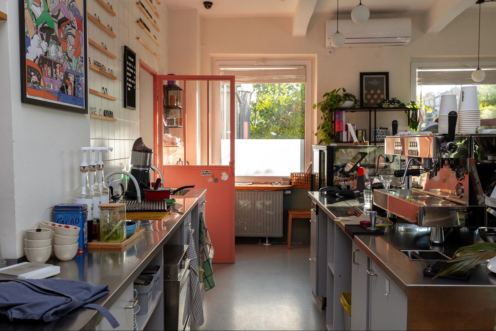Wnętrze kawiarni z dwoma stanowiskami roboczymi, wyposażonymi w ekspresy do kawy, zlewozmywaki i akcesoria baristyczne, z widoczną otwartą, pomarańczową przegrodą prowadzącą do zaplecza.