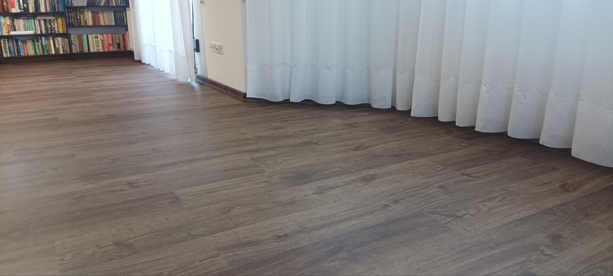 Ułożone panele podłogowe w pokoju z książkami i białymi zasłonami z subtelnym haftem.