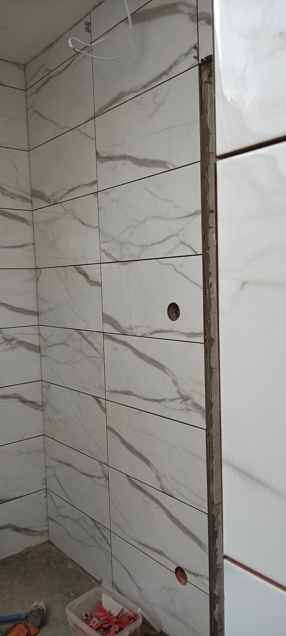 Ściana łazienki wyłożona białymi płytkami imitującymi marmur, z widocznym otworem na gniazdko i narzędziami glazurniczymi w dolnej części kadru. Widoczny fragment sufitu.
