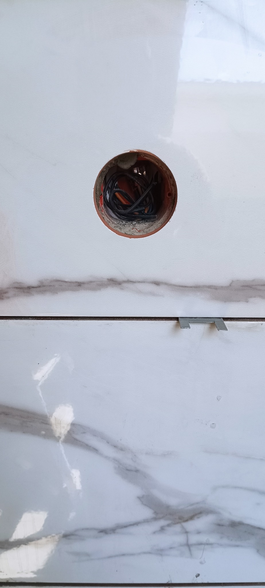 Białe płytki imitujące marmur z otworem na puszkę elektryczną, w której widoczne są przewody. Klipsy dystansowe między płytkami.