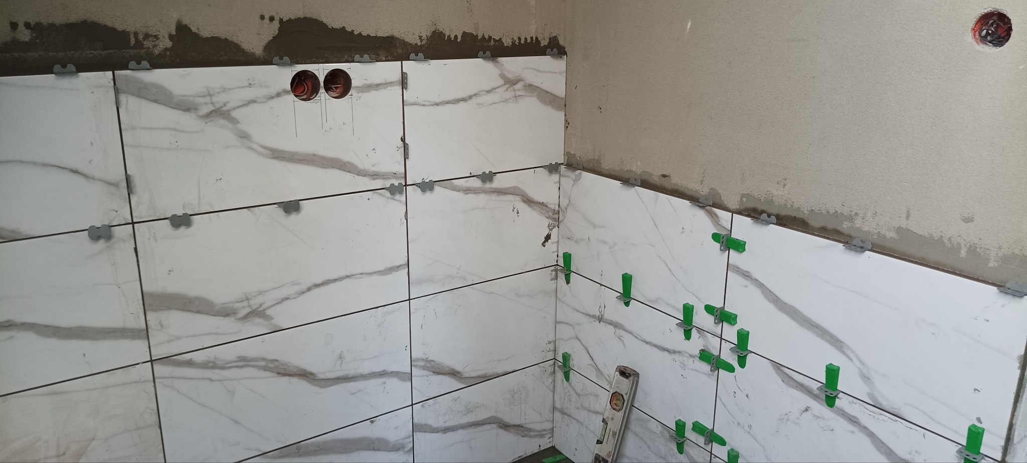 Białe płytki imitujące marmur, ułożone na ścianie w rogu pomieszczenia, z użyciem klipsów poziomujących i poziomicy. Widoczne puszki elektryczne.