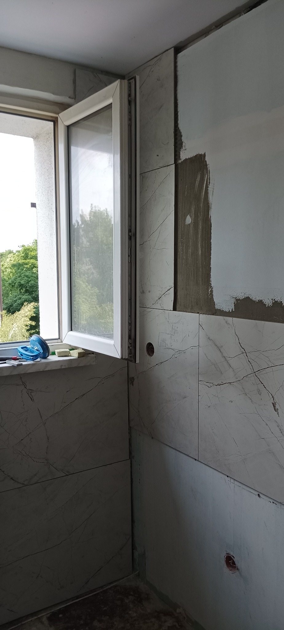 Fragment łazienki w trakcie wykończenia: marmurowe płytki na ścianach, otwarte okno z widokiem na zieleń, ślady po szpachlowaniu i gniazdko elektryczne.